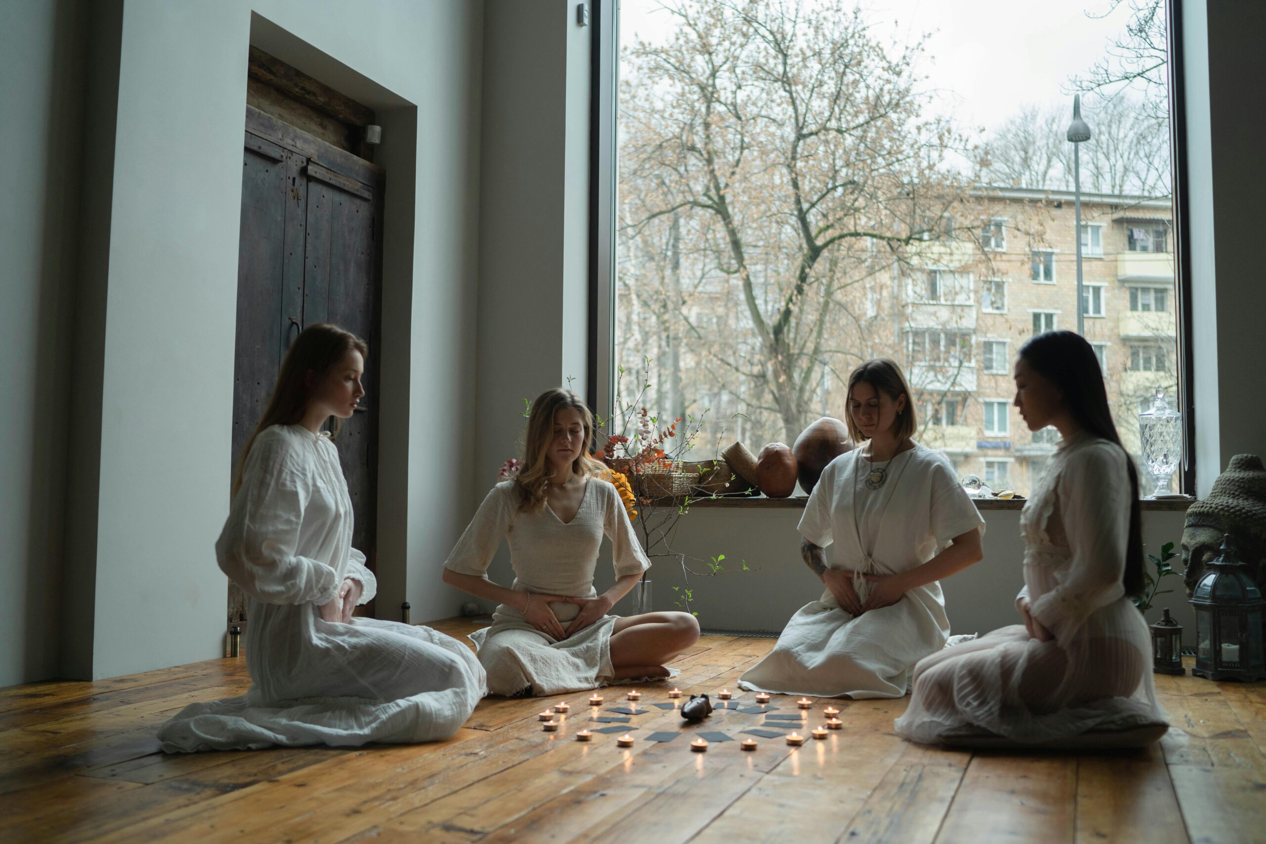 Four women in white dresses meditate with candles on a wooden floor indoors.